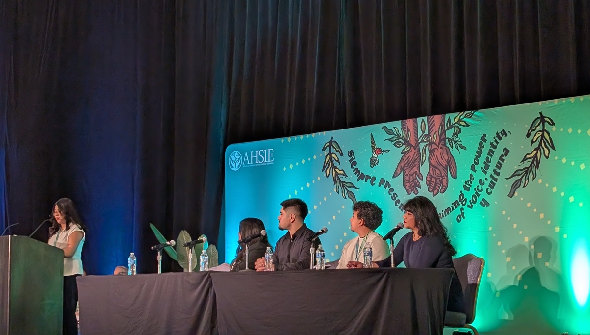 Leslie Arias stands at a podium in front of a panel of speakers